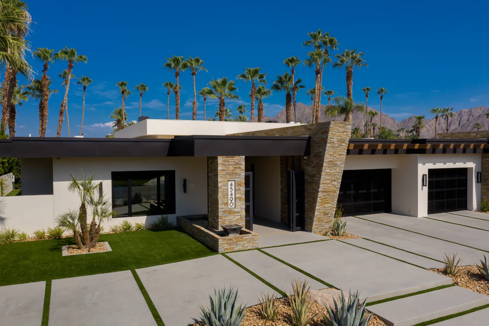 Single-storey desert modern home with stacked stone entry columns, glass garage doors and a palm-lined driveway